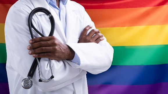 A Black male physician in white coat with arms crossed and holding a stethoscope in front of the LGBTQ flag.