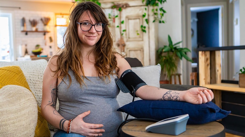 A pregnant woman taking her blood pressure at home and smiling at the camera person.