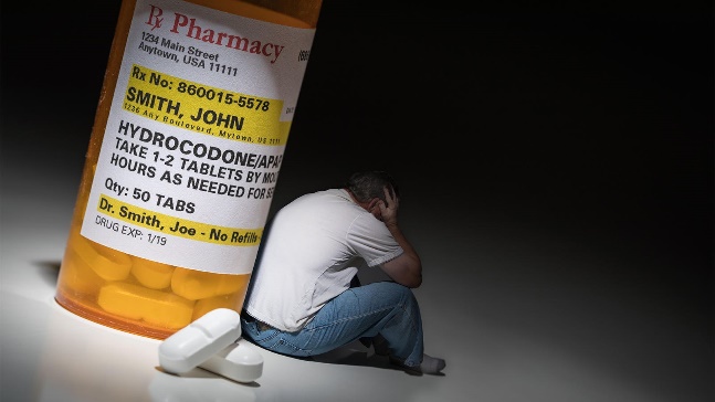 A photo composite of a man with his head in his hands leaning against a large prescription bottle of opioids.