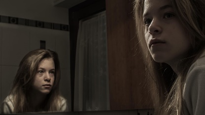 A photo of a young woman looking away from a mirror while her reflection looks at her.