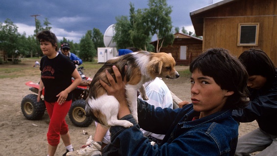 A photo of a teen boy holding up his dog with his family in the background in Alaska.